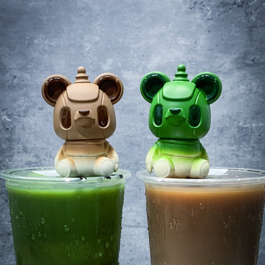 Two bear-shaped drink covers, one beige and one green, on cups of green and brown beverages against a gray background.