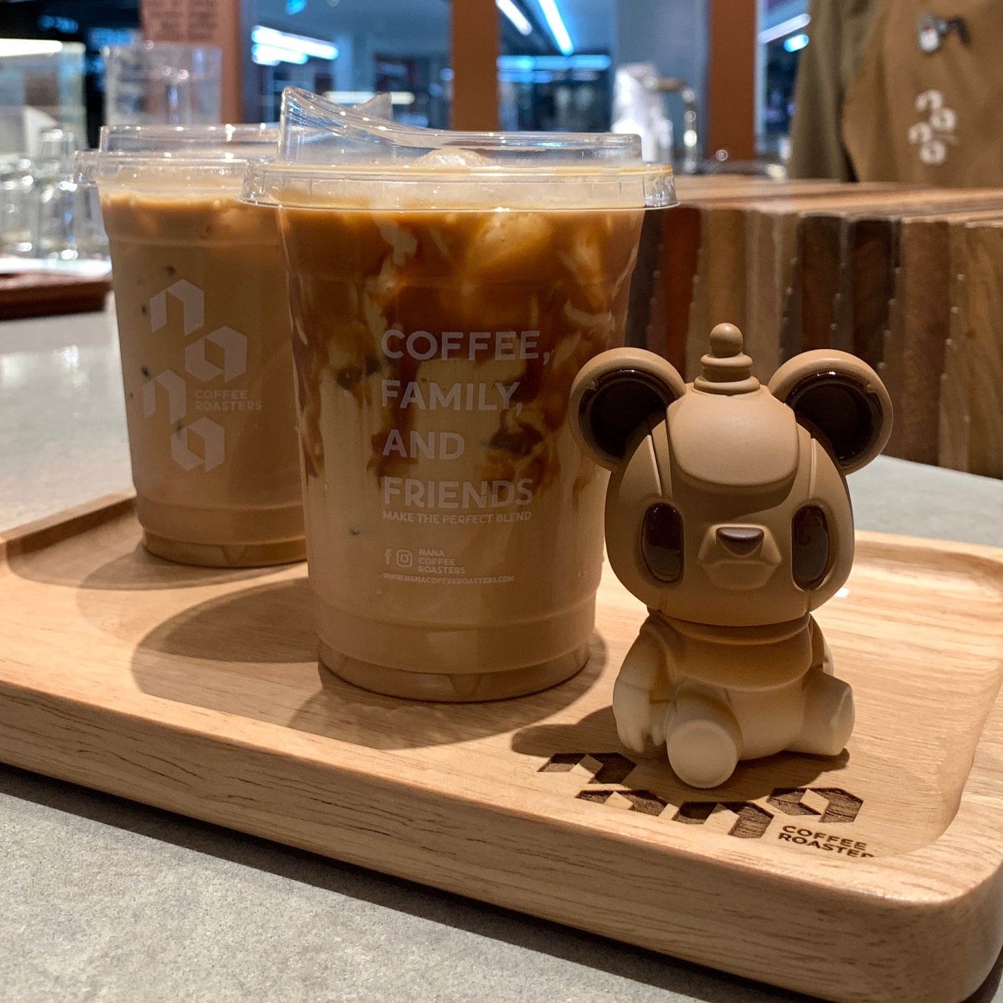 Two iced coffee drinks on a tray with a small bear figurine in a coffee shop setting.