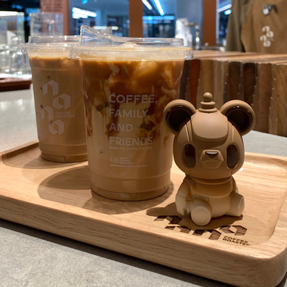 Two iced coffee drinks on a tray with a small bear figurine in a coffee shop setting.